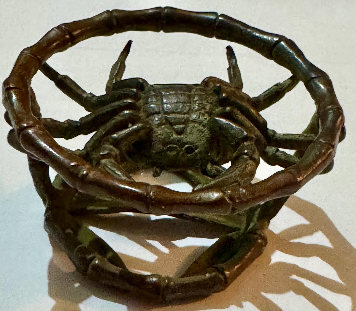 Bronze crab figurine with raised claws inside bamboo-form cage, photographed against white background with tan cardboard base