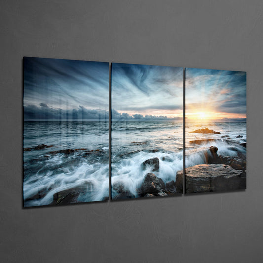 Three-panel glass wall art displaying a coastal sunset scene with waves crashing over dark rocks and golden sky.