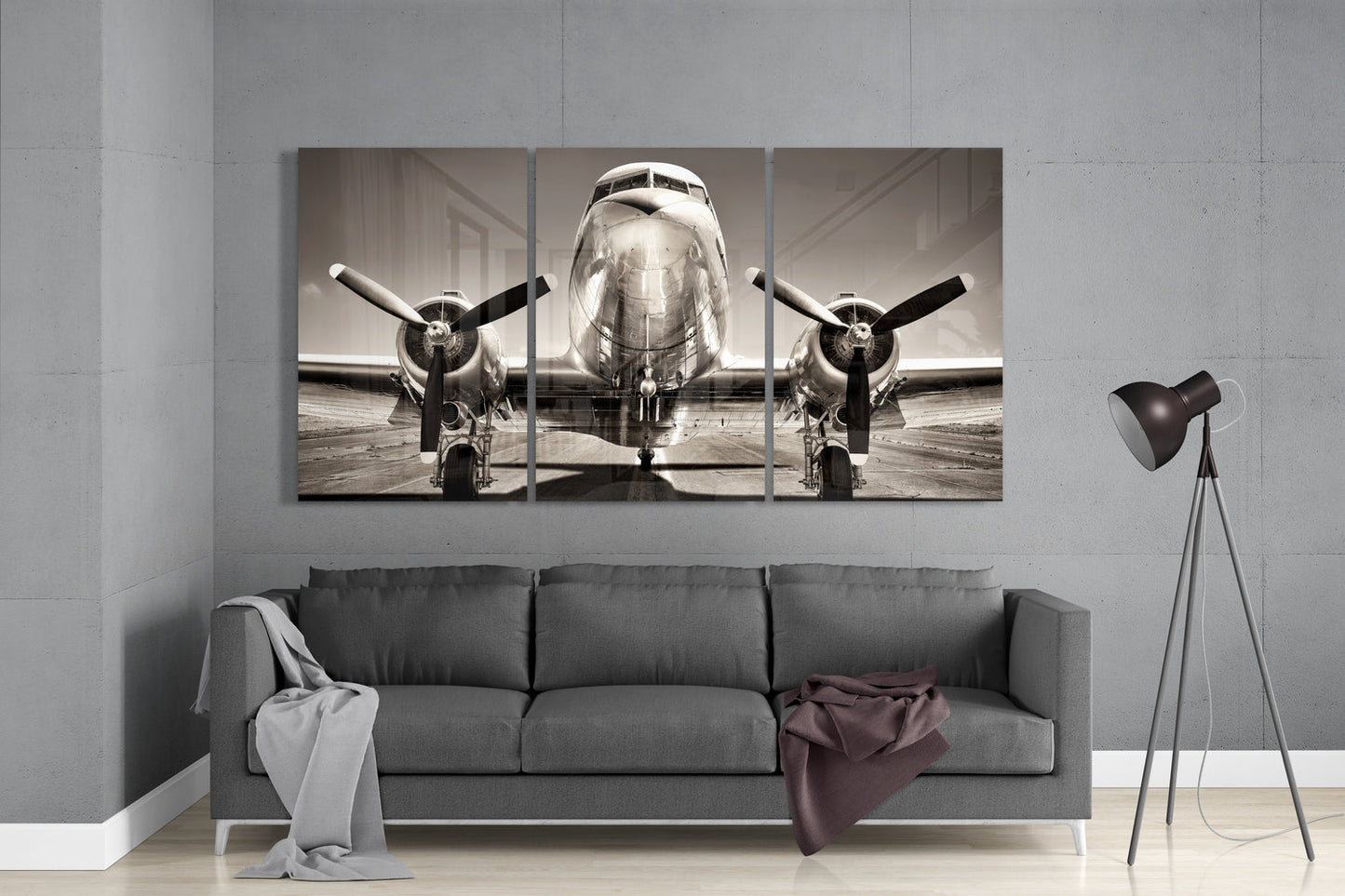 Three-panel sepia-toned glass artwork displaying front view of vintage twin-engine aircraft on tarmac, installed above gray sofa in modern interior.