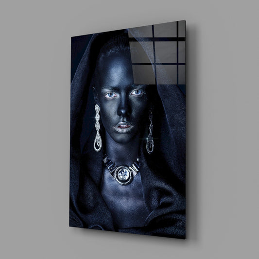 Glass wall art mounted on gray wall showing woman with dark skin, striking blue eyes, silver jewelry, and dark draped fabric against dark background.