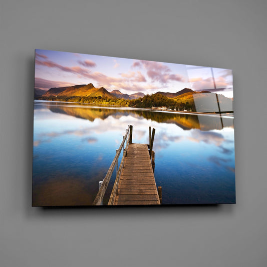 Wooden jetty extending into calm lake with golden-lit mountains and forest reflected in water, displayed as glass wall art on gray background.