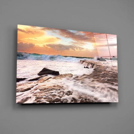 Coastal sunset scene with crashing waves and rocky shoreline printed on glass panel mounted on gray wall.