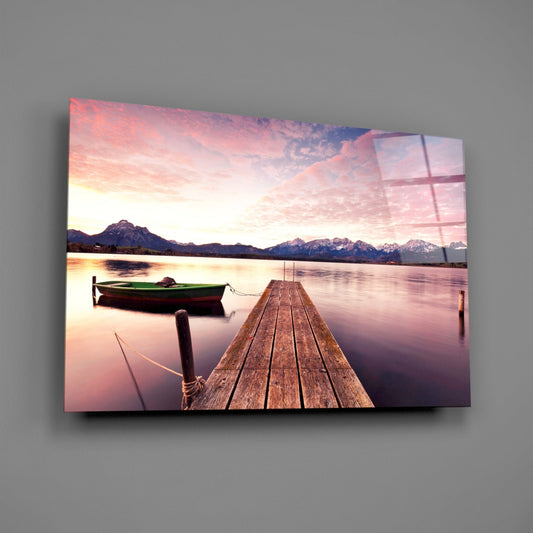 Wooden pier extending into calm pink-toned lake with green boat, snow-capped mountains, and sunset sky on glass wall art panel.