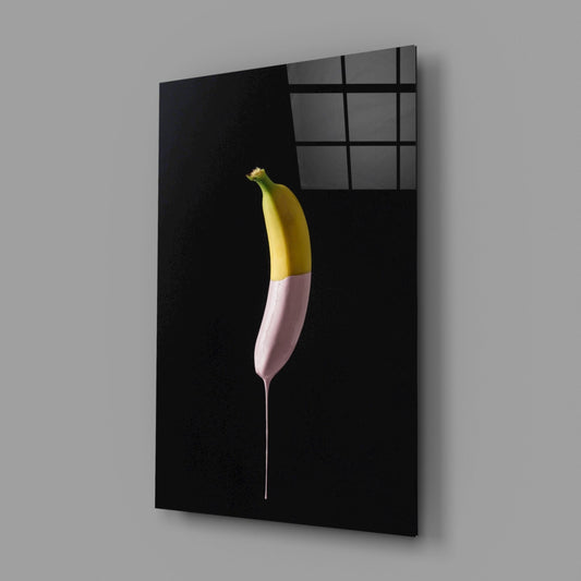 A banana with yellow top and pink-dipped bottom section mounted on black glass against a dark interior wall with gridded window visible above.
