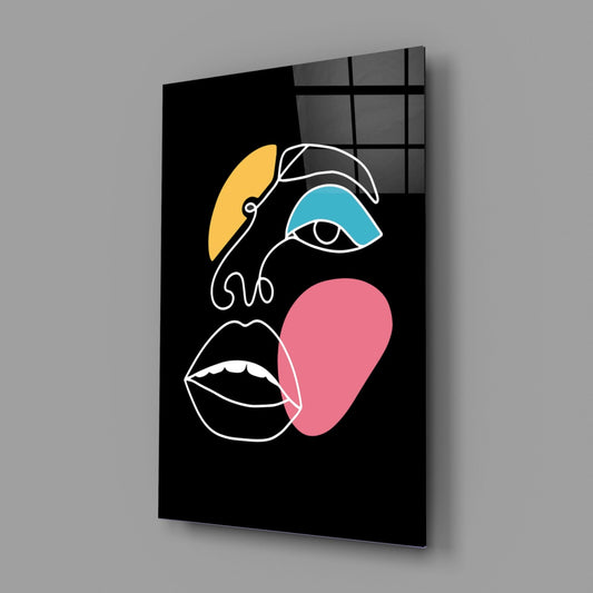 Modern glass wall art panel displaying an abstract face with white line-drawn features, yellow crescent, teal eye patch, and pink chin area on black background.