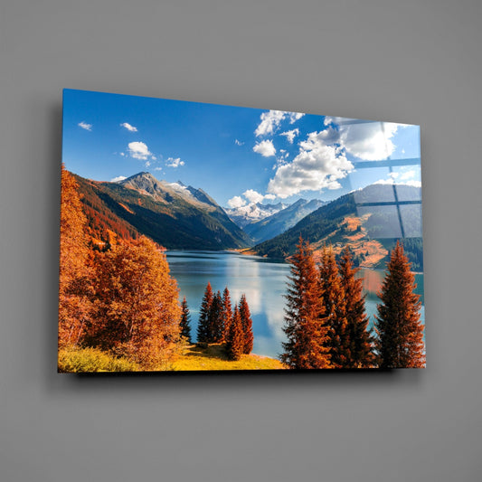 Framed glass wall art displaying an autumn mountain lake valley with golden larch trees, snow-capped peaks, and blue sky with clouds.
