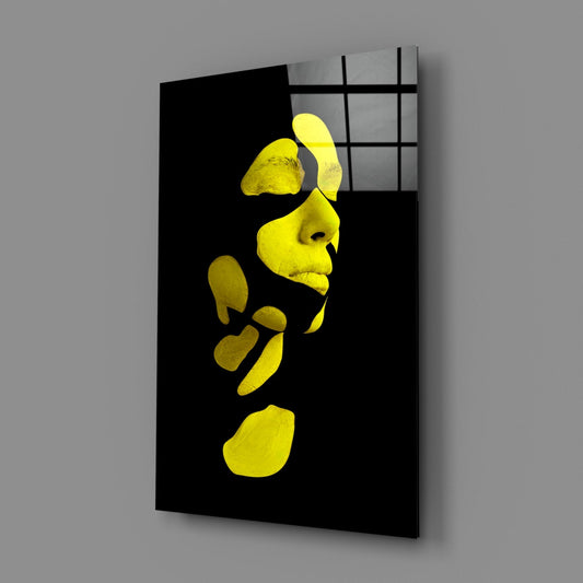 Angled perspective of black glass wall art displaying fragmented yellow face and hand forms against dark background with gridded window visible above.