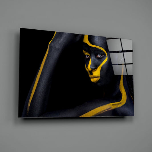 Glass wall art displaying a woman's face with black and gold paint stripes, mounted at an angle against a gray wall.