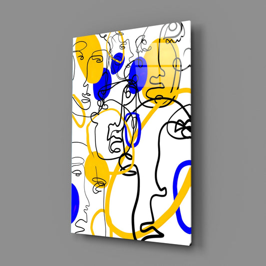Angled view of glass wall art displaying continuous line-drawn faces in black with yellow and blue geometric fills on white background.