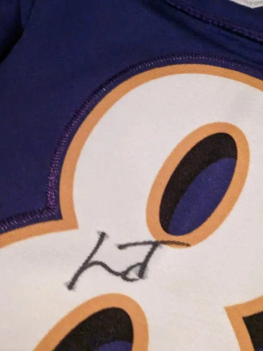 Close-up of a purple and white jersey with an orange-outlined white number and a black handwritten signature.