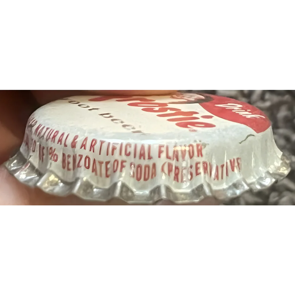 Angled side view of a Frostie root beer cap showing red printed skirt text about natural and artificial flavor and benzoate of soda, with part of the red 'Drink' area.