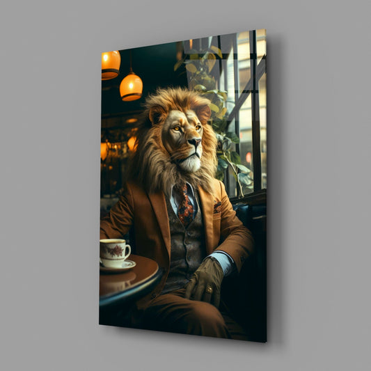 Lion in brown blazer and tie seated at café table with coffee cup, hanging orange lamps and plants visible behind, displayed as glass wall art panel.