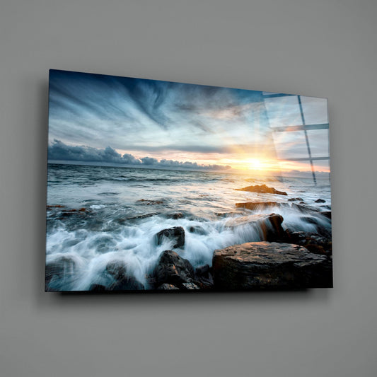 Coastal sunset scene with waves crashing over dark rocks printed on glass wall art mounted on gray wall.