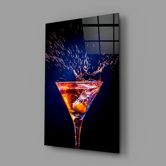 Angled perspective view of glass wall art panel displaying a martini glass with amber liquid and dynamic splash against dark blue background, mounted on gray wall.