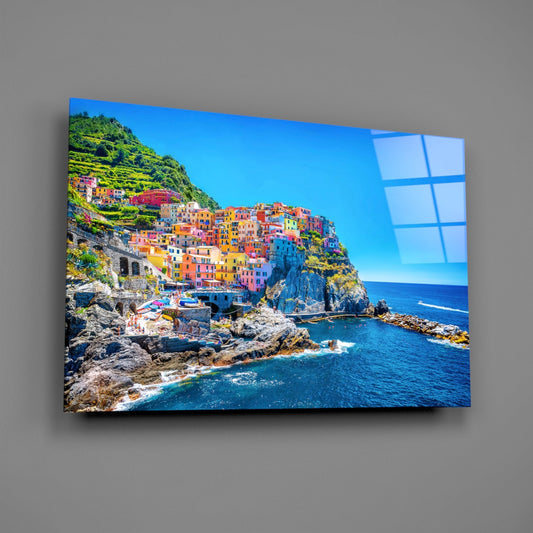 Colorful cliffside village with blue sea reflected on glass wall art mounted on gray wall, angled perspective view.