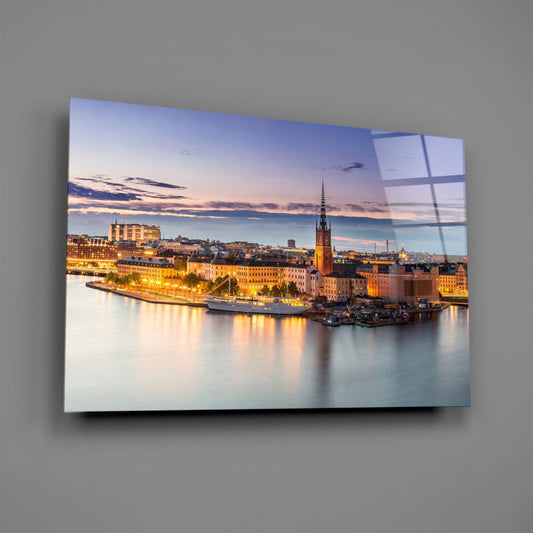 Glass wall art mounted on gray wall showing Stockholm waterfront at twilight with illuminated buildings, church spire, and water reflections.