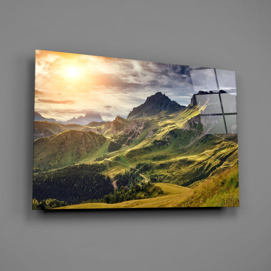Glass wall art mounted on gray wall displays alpine valley landscape with green slopes, forested areas, and distant mountains under golden sunset light.