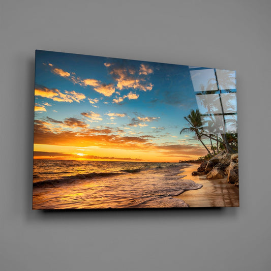 Beach sunset glass wall art mounted on gray wall, showing tropical shore with golden sky, ocean waves, and palm trees reflected in glass panel.