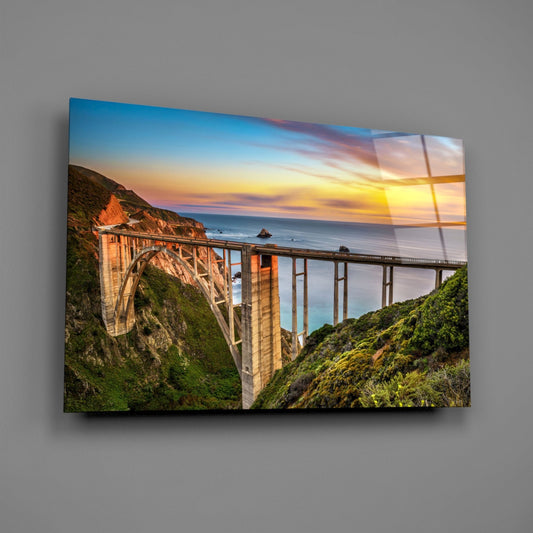 Bixby Creek Bridge glass wall art displayed at an angle on gray wall, showing sunset coastal landscape with bridge and green cliffs.