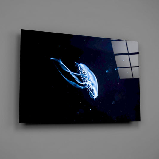 Glass wall art panel mounted on gray wall showing bioluminescent jellyfish glowing blue against black starry background.