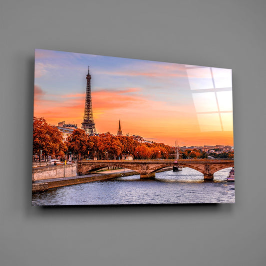 Glass wall art displaying Paris sunset with Eiffel Tower, Seine River bridge, and autumn trees in warm orange and golden tones.