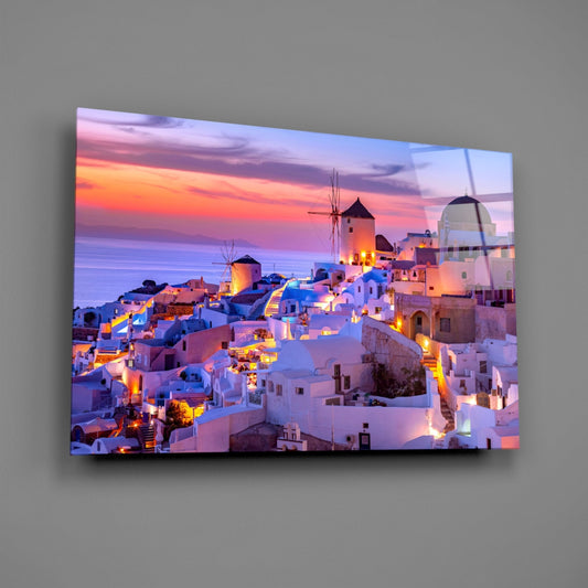Glass wall art displaying Santorini's whitewashed buildings and windmills at sunset, mounted on gray wall showing vibrant purple and orange sky.