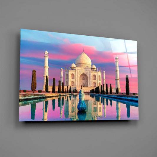 Glass wall art displaying the Taj Mahal with pink and blue sunset sky, cypress trees, and reflective water canal, mounted on gray wall.
