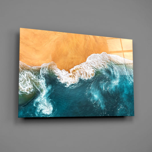 Aerial view of amber sand beach meeting turquoise ocean waves with white foam, printed on glass panel mounted on gray wall.