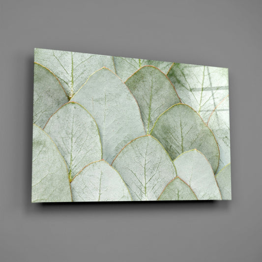 Angled front view of glass wall art featuring layered sage and cream eucalyptus leaves with visible veining against gray background.