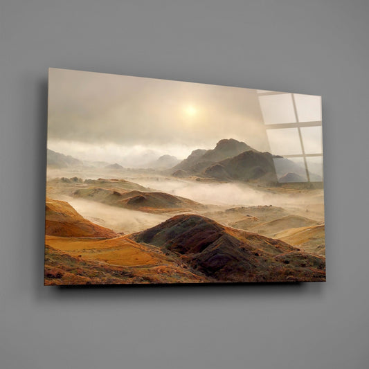 Glass wall art displaying layered desert hills with golden-brown tones and white fog, mounted on gray wall with window-pane reflection overlay.