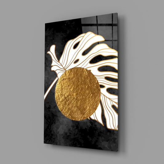 Angled perspective of glass wall art displaying white line-drawn monstera leaf with textured gold circle center against black background.