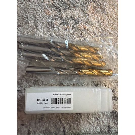 Four gold and silver coated drill bits in clear plastic packaging next to Haas Tooling label on concrete surface.