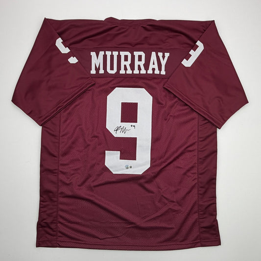 Flat-lay overhead view of maroon college football jersey with "MURRAY" nameplate and number 9 in white, featuring visible autograph on the jersey.