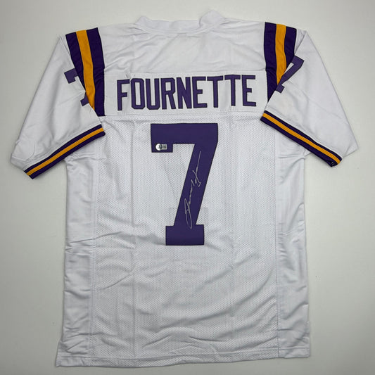 White LSU football jersey displayed flat, showing back with "FOURNETTE" nameplate, number 7, purple and gold shoulder stripes, and autograph on number.