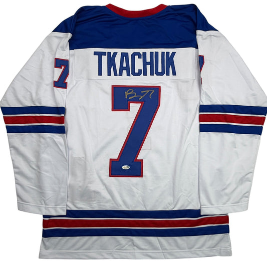 Back view of white hockey jersey with navy and red stripes, displaying "TKACHUK" and number 7 with autograph.