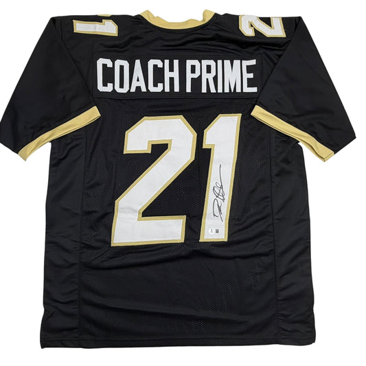 Black college football jersey with gold and white trim, "COACH PRIME" across chest, number 21 with signature, flat lay front view.