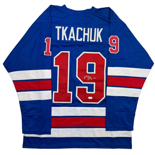 Back view of blue hockey jersey with red number 19, white "TKACHUK" nameplate, and red-white-blue horizontal stripes on sleeves.