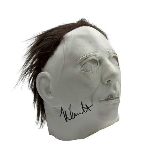 White Michael Myers mask with dark brown hair on left side, shown in three-quarter view with handwritten signature on cheek.