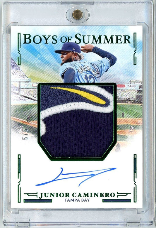 Baseball card featuring Junior Caminero in pitching pose with navy jersey, dark fabric relic patch, and blue autograph on white background with green borders.