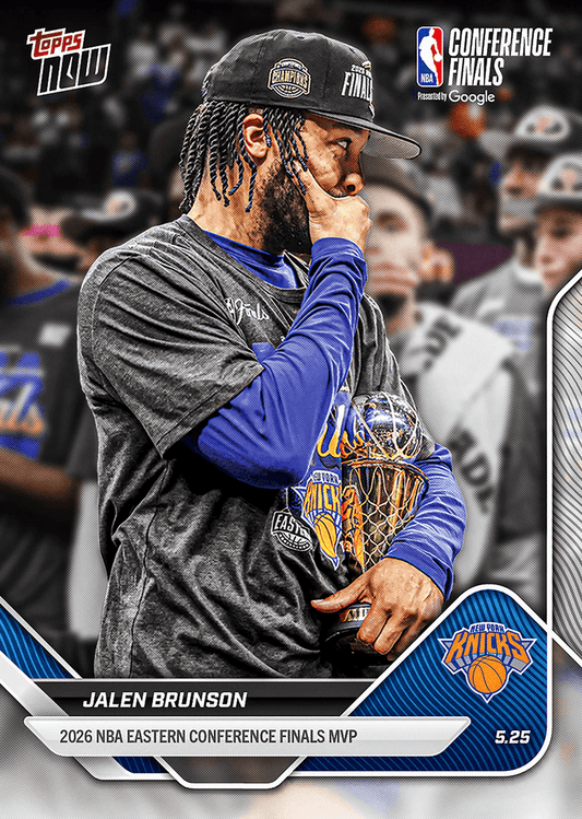 Jalen Brunson wearing black cap and blue Knicks warmup gear, holding trophy while touching his face, photographed in profile during celebration.