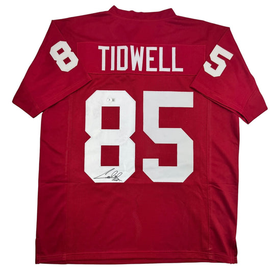 Red football jersey with white "TIDWELL" nameplate and "85" number, displaying autograph on front, flat lay view.