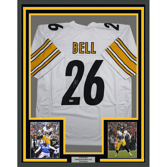 Framed white Pittsburgh Steelers jersey #26 signed by Le'Veon Bell, displayed with yellow and black borders and two action photographs below.