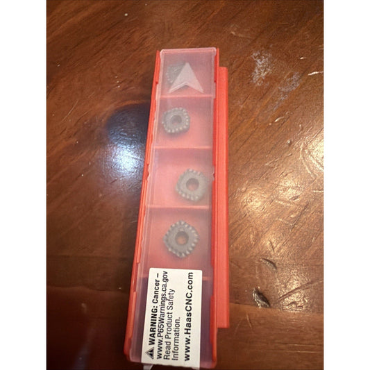 Red plastic storage case containing five gray carbide inserts with warning label visible on wooden surface.