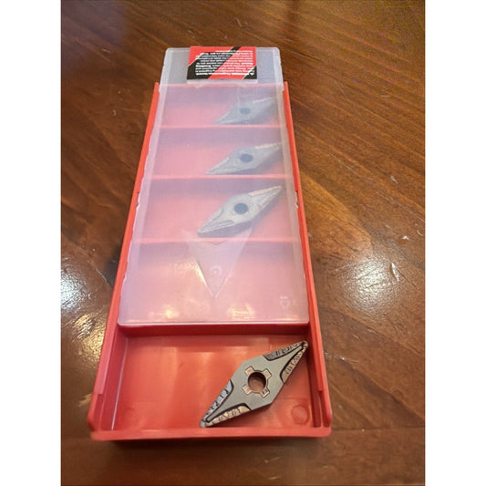 Red plastic organizer box with compartments displaying three triangular carbide inserts and one loose insert at bottom, wooden surface background.