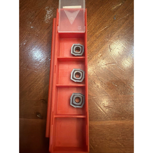 Red plastic storage case with transparent hinged lid containing three metallic square carbide inserts arranged vertically, viewed from above on wooden surface.