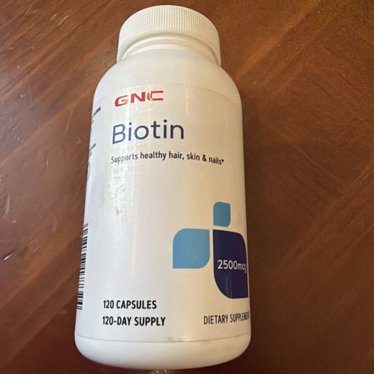 GNC Biotin 2500mcg supplement bottle with white cap on wooden surface, front label visible showing product name and 120 capsules.