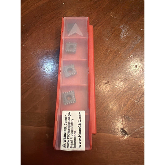 Red plastic compartment box containing five gray carbide inserts with warning label visible on wooden surface, top-down view.