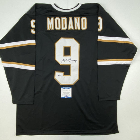 Back view of black Dallas hockey jersey number 9 with MODANO nameplate, gold and white trim, displaying signature and Beckett authentication sticker.