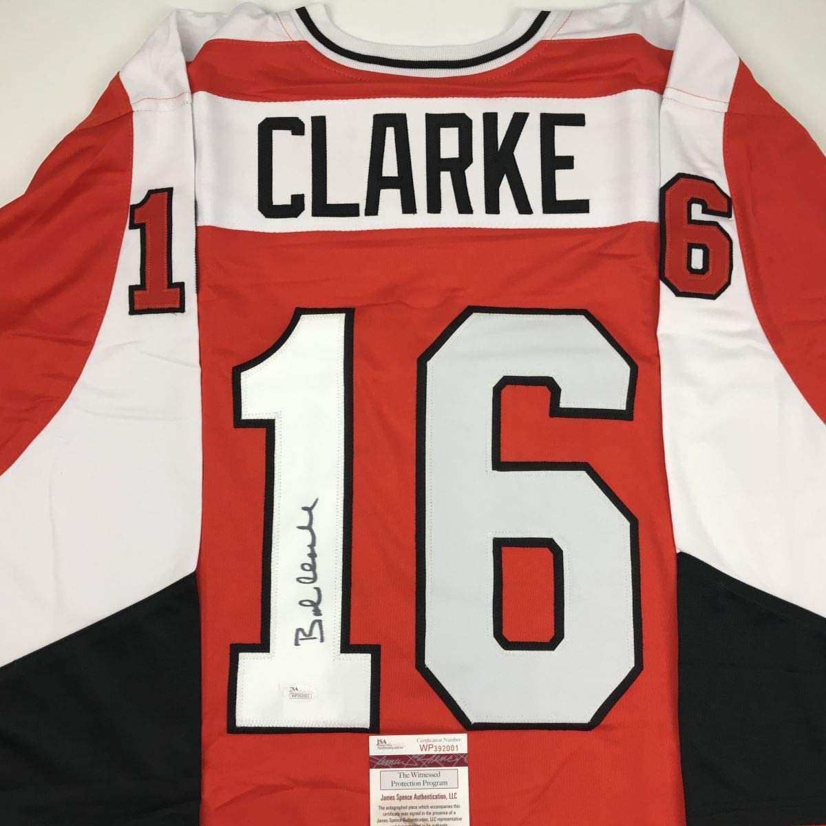 Red and white Philadelphia hockey jersey number 16 with "CLARKE" nameplate, signed in black ink, displayed flat with JSA authentication label visible at bottom.