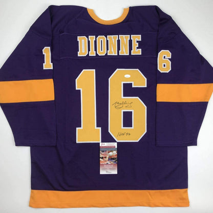 Flat lay of signed purple and gold LA Kings hockey jersey with number 16, "DIONNE" nameplate, and JSA authentication certificate attached.